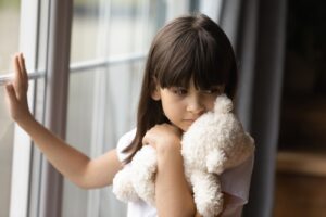 A young girl with straight dark hair stands by a window, holding a white teddy bear tightly to her chest. She looks out the window with a sad and distant expression, suggesting feelings of loneliness or distress.
