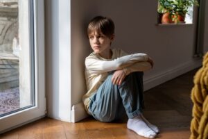 A young boy in a long-sleeve cream shirt and blue pants sits curled up on the floor by a large window. He leans against the wall with a solemn, contemplative expression, his arms resting on his knees, as sunlight filters into the room.